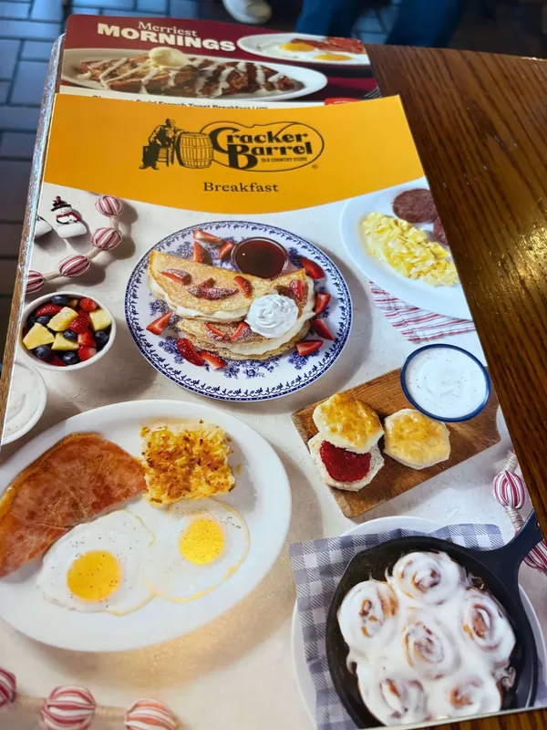 Cracker Barrel breakfast menu cover on a wooden table, with the barrel-and-man logo in a yellow band over photographs of pancakes, eggs, biscuits and cinnamon rolls.