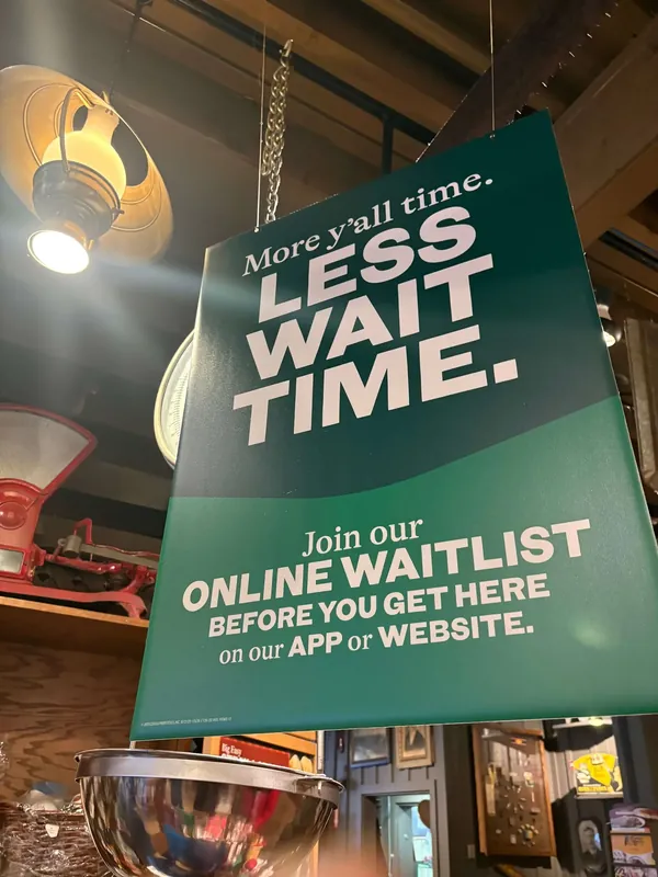 Green hanging sign in the store reading More y'all time, Less wait time, promoting the online waitlist, above shelves of country store goods.