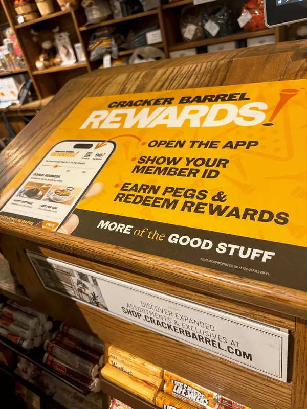 Yellow Cracker Barrel Rewards sign on a wooden retail counter, showing a phone with the app and three steps to earn and redeem pegs.