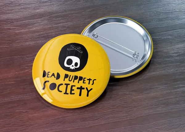 Yellow enamel pin on a dark wood surface carrying the Dead Puppets Society skull and lettering, with a second pin face down behind it.
