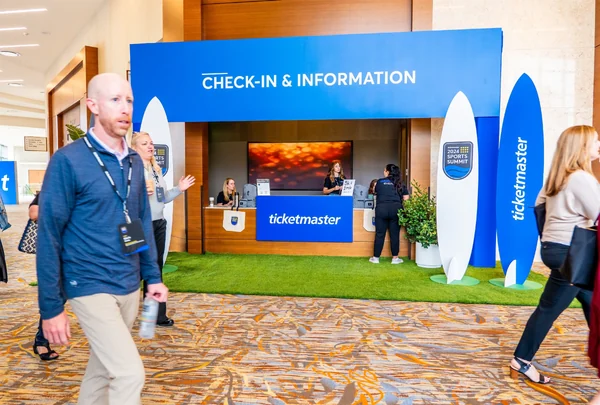 Branded check-in and information desk at the conference, with a blue backdrop, surfboard-shaped Ticketmaster signs, and attendees walking past.