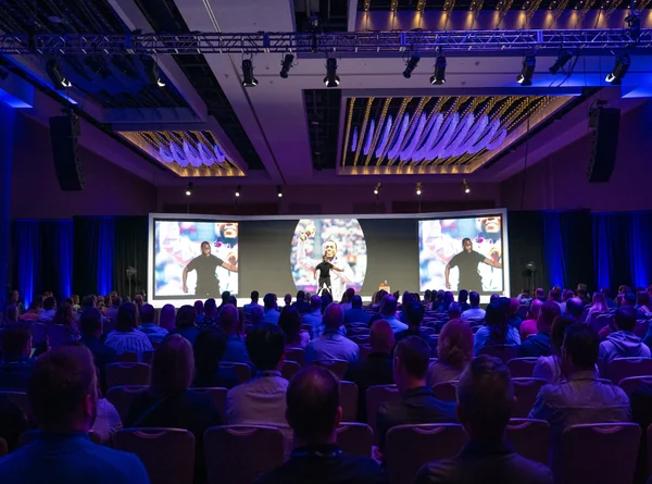 Darkened ballroom during a keynote, with attendees seated in rows facing a wide stage and three lit screens.