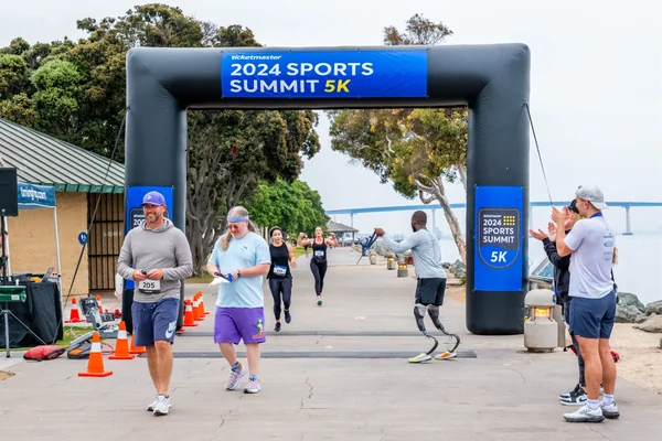 Runners passing under an inflatable 2024 Sports Summit 5K arch on a tree-lined path.