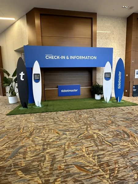 Check-in and information desk with a blue backdrop, two surfboard-shaped signs, and artificial turf at its base.