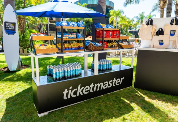 Outdoor refreshment station on a lawn, with a white umbrella over a black Ticketmaster-branded counter stocked with bottled drinks.